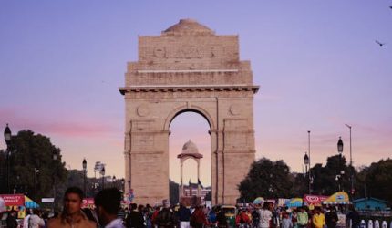 A bustling scene at India Gate with a vibrant crowd under a colorful sunset sky.